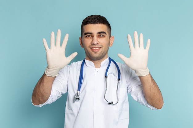 Vista frontal del joven médico en traje blanco con estetoscopio azul sonriendo y mostrando las manos