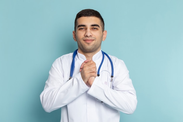 Vista frontal del joven médico en traje blanco con estetoscopio azul sonriendo posando