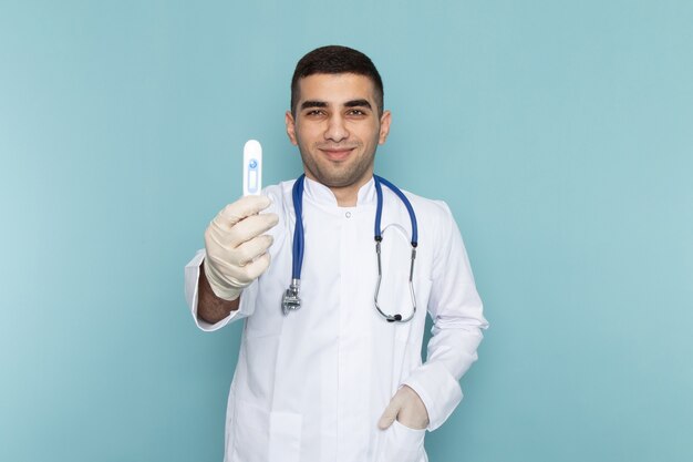 Vista frontal del joven médico en traje blanco con estetoscopio azul sonriendo y sosteniendo el dispositivo