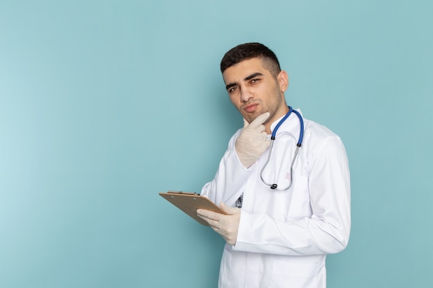 Vista frontal del joven médico en traje blanco con estetoscopio azul sosteniendo el bloc de notas con expresión de pensamiento