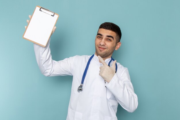 Vista frontal del joven médico en traje blanco con estetoscopio azul sosteniendo el bloc de notas con una sonrisa