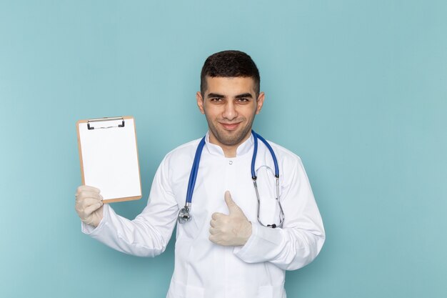 Vista frontal del joven médico en traje blanco con estetoscopio azul sosteniendo el bloc de notas con una sonrisa