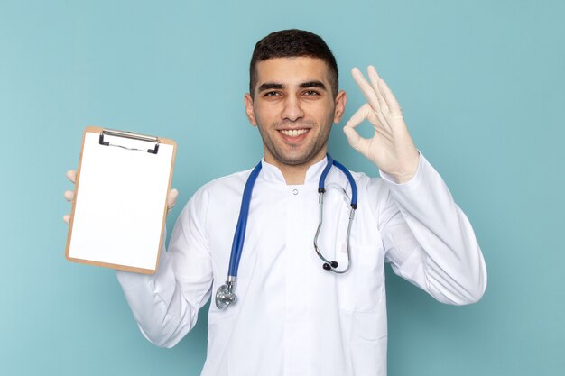 Vista frontal del joven médico en traje blanco con estetoscopio azul sosteniendo el bloc de notas con una sonrisa
