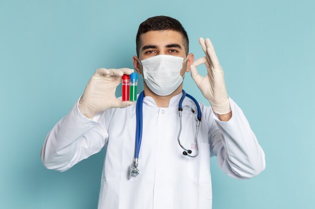 Vista frontal del joven médico en traje blanco con estetoscopio azul sosteniendo frascos con máscara