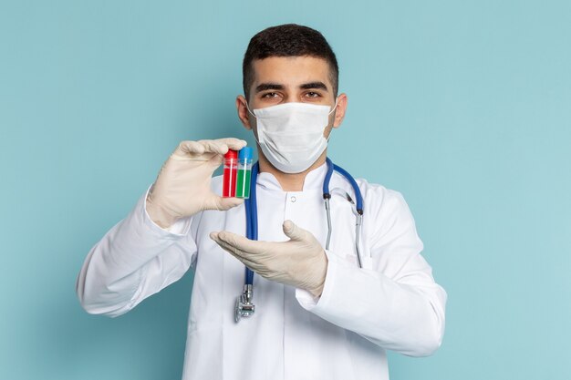 Vista frontal del joven médico en traje blanco con estetoscopio azul sosteniendo frascos con máscara