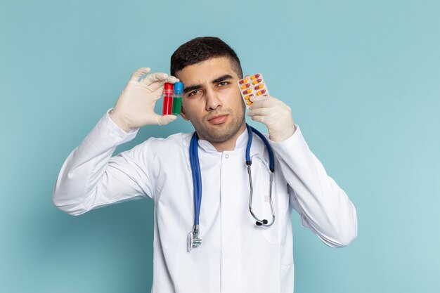 Vista frontal del joven médico en traje blanco con estetoscopio azul sosteniendo frascos y pastillas