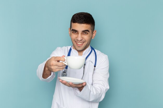 Vista frontal del joven médico en traje blanco con estetoscopio azul sosteniendo una taza de café con una sonrisa