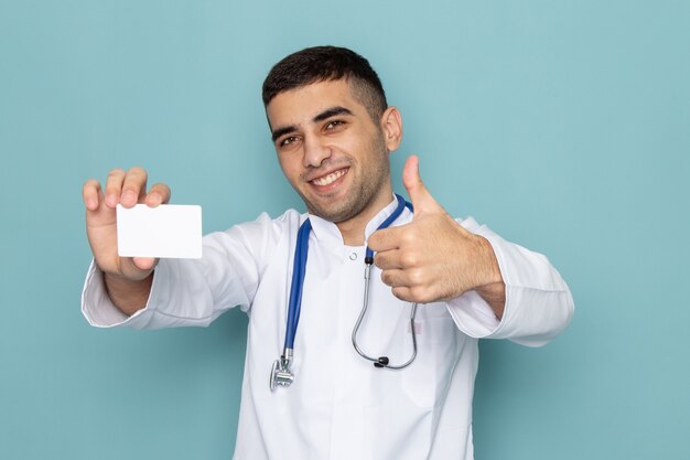 Vista frontal del joven médico en traje blanco con estetoscopio azul con tarjeta blanca