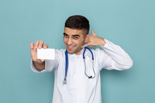 Vista frontal del joven médico en traje blanco con estetoscopio azul con tarjeta blanca