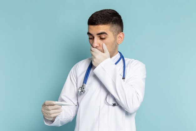 Vista frontal del joven médico en traje blanco con estetoscopio azul con termómetro con expresión de sorpresa en el trabajo de trabajo de escritorio azul medicina hospital médico