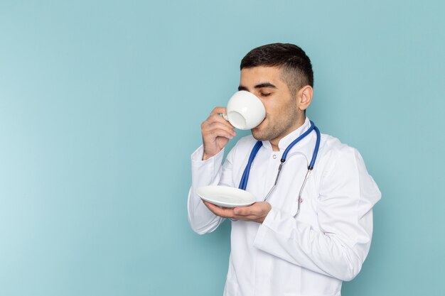 Vista frontal del joven médico en traje blanco con estetoscopio azul tomando café