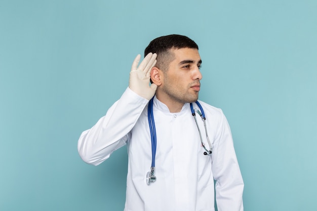 Vista frontal del joven médico en traje blanco con estetoscopio azul tratando de escuchar