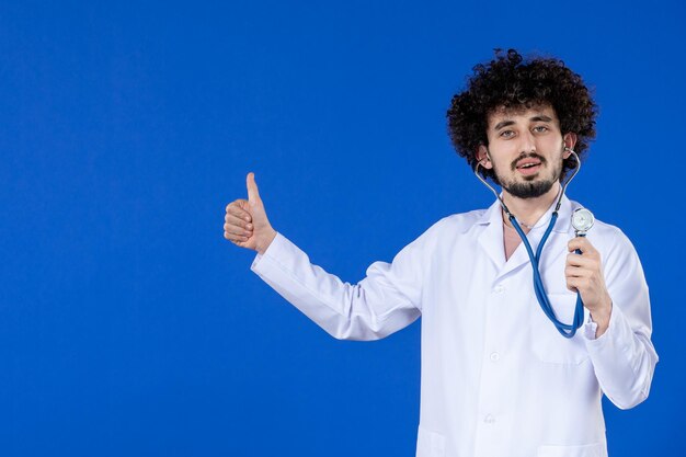Vista frontal del joven médico en traje médico con estetoscopio sobre superficie azul