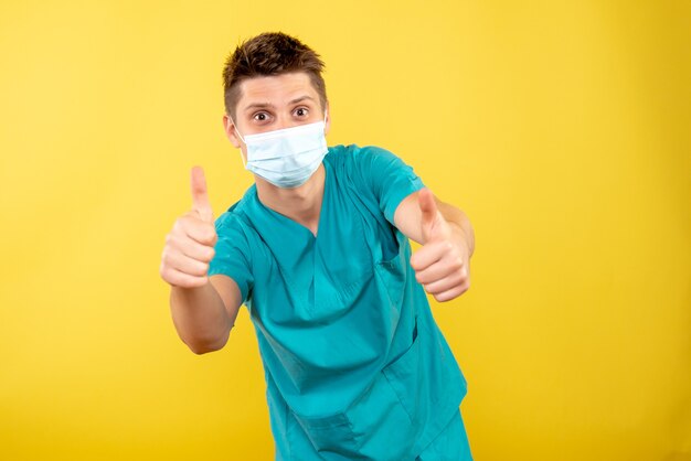 Vista frontal joven médico en traje médico con máscara estéril sobre fondo amarillo