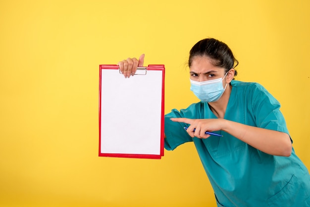 Vista frontal del joven médico en uniforme apuntando al portapapeles en la pared amarilla