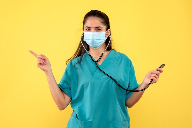 Vista frontal joven médico en uniforme apuntando a algo de pie sobre fondo amarillo