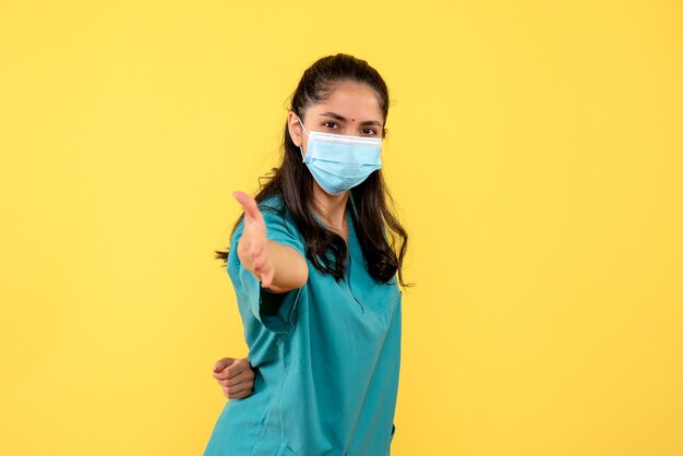 Vista frontal joven médico en uniforme poniendo una mano detrás de su espalda de pie sobre fondo amarillo