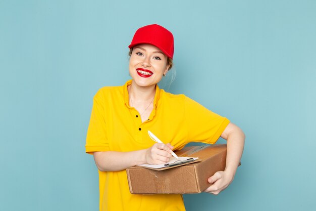 Vista frontal joven mensajero en camisa amarilla y capa roja con bloc de notas de paquete sonriendo en el trabajo de espacio azul