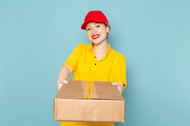 Vista frontal joven mensajero con camisa amarilla y capa roja sonriendo y sosteniendo el paquete en el trabajo del espacio azul