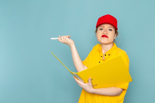 Vista frontal joven mensajero con camisa amarilla y capa roja sosteniendo un archivo amarillo escribiendo notas sobre el trabajador espacial azul