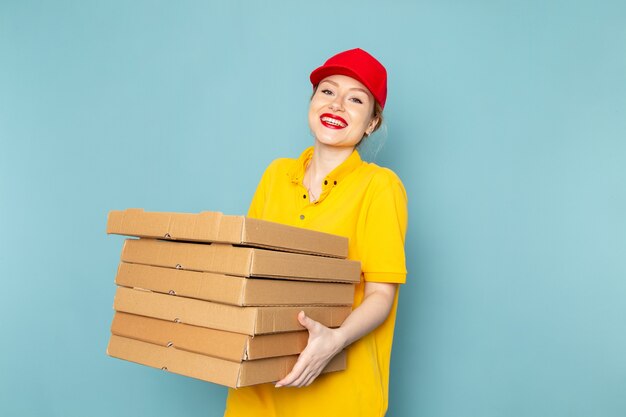Vista frontal joven mensajero con camisa amarilla y capa roja sosteniendo paquetes con una sonrisa en el trabajo del espacio azul