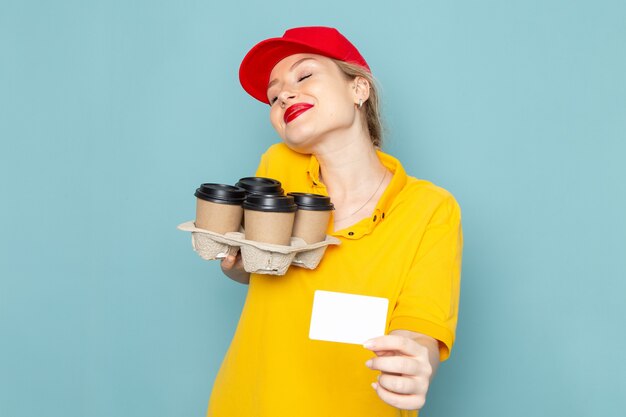 Vista frontal joven mensajero con camisa amarilla y capa roja sosteniendo tazas de café de plástico tarjeta blanca con sonrisa en el espacio azul