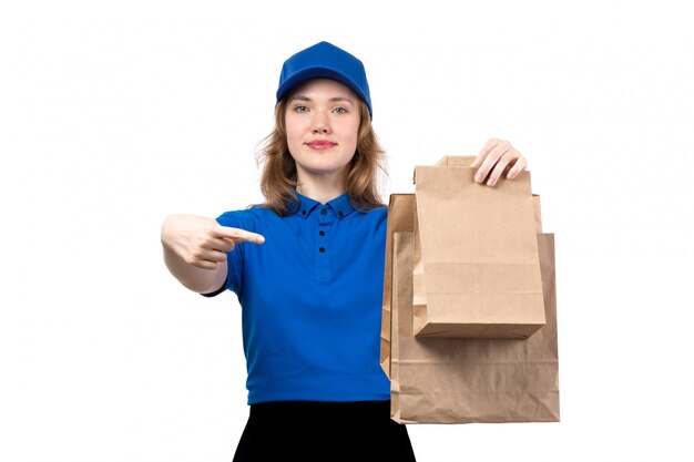 Una vista frontal joven mensajero en camisa azul gorra azul y pantalón negro sonriendo sosteniendo paquetes de alimentos en blanco