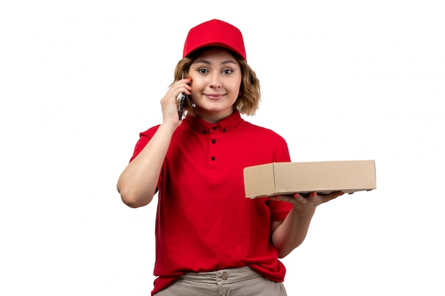 Una vista frontal joven mensajero en camisa roja gorra roja con caja de entrega sonriendo hablando por teléfono