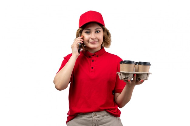 Una vista frontal joven mensajero en camisa roja gorra roja sosteniendo tazas de café hablando por teléfono sonriendo