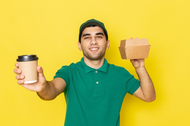 Vista frontal joven mensajero en camisa verde gorra verde con paquete de entrega y taza de café sonriendo en amarillo