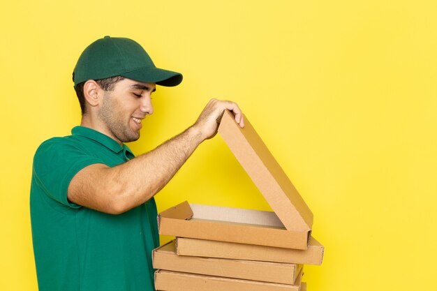 Vista frontal joven mensajero en camisa verde gorra verde sonriendo y abriendo cajas de entrega en el color de fondo amarillo trabajo entregando servicio