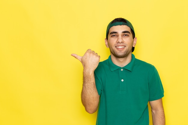 Vista frontal joven mensajero en camisa verde gorra verde sonriendo y señalando con el dedo sobre el fondo amarillo trabajo entregando color de servicio