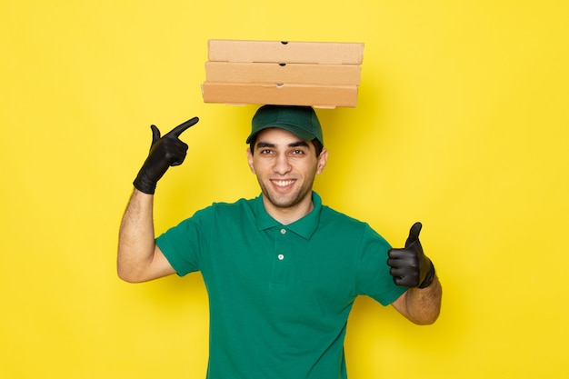 Vista frontal joven mensajero en camisa verde gorra verde sosteniendo cajas de entrega en la cabeza sonriendo en amarillo