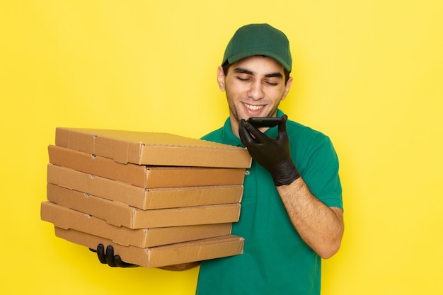 Vista frontal joven mensajero en camisa verde gorra verde sosteniendo cajas de entrega y hablando por teléfono sonriendo en amarillo