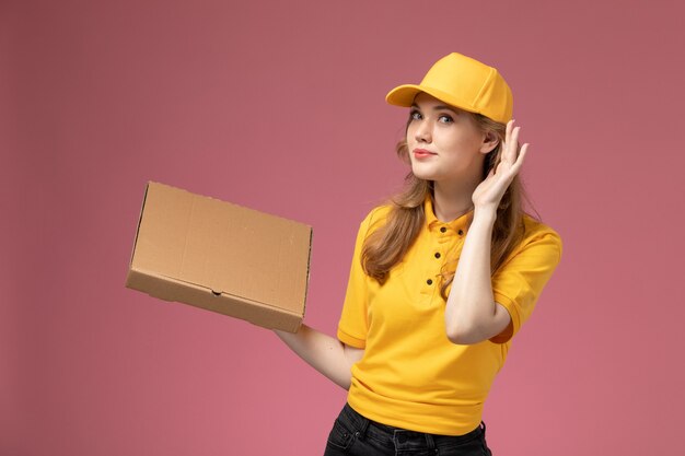 Vista frontal joven mensajero en capa amarilla uniforme amarillo con caja de entrega de alimentos en el servicio de trabajo de entrega uniforme de fondo rosa oscuro