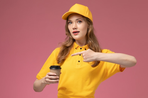 Vista frontal joven mensajero en capa amarilla uniforme amarillo sosteniendo la taza de café en el fondo rosa oscuro servicio de entrega uniforme trabajadora