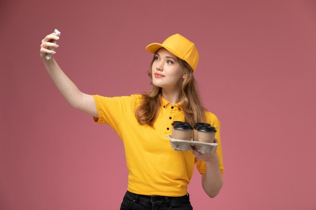 Vista frontal joven mensajero en capa amarilla uniforme amarillo tomando un selfie con café en el color de servicio de trabajo de entrega uniforme de fondo rosa oscuro