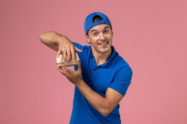 Vista frontal joven mensajero en capa uniforme azul con paquete de comida de entrega en la pared rosa