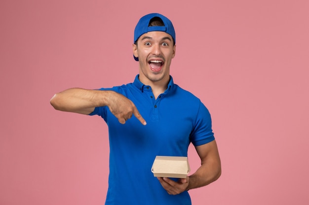 Vista frontal joven mensajero en capa uniforme azul con paquete de comida de entrega en la pared rosa