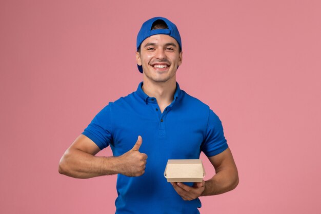 Vista frontal joven mensajero en capa uniforme azul con paquete de comida de entrega en la pared rosa