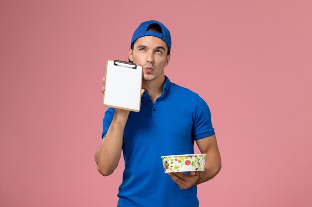 Vista frontal joven mensajero en capa uniforme azul sosteniendo el bloc de notas y cuenco de entrega redonda pensando en la pared rosa