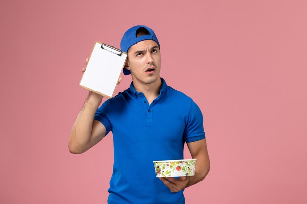 Vista frontal joven mensajero en capa uniforme azul sosteniendo el bloc de notas y el tazón de entrega redondo pensando en la pared rosa