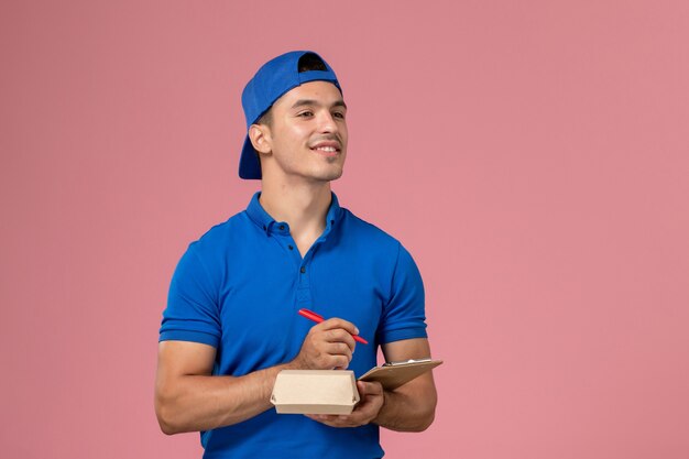 Vista frontal joven mensajero con capa uniforme azul sosteniendo un pequeño paquete de comida de entrega y un bloc de notas escribiendo notas en la pared rosa claro