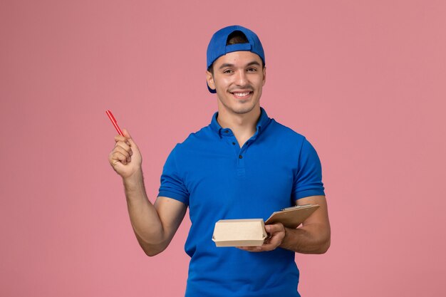 Vista frontal joven mensajero con capa uniforme azul sosteniendo un pequeño paquete de comida de entrega y un bloc de notas escribiendo notas en la pared rosa claro