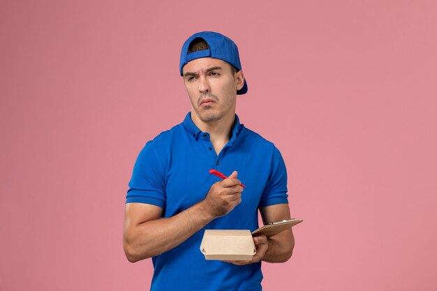 Vista frontal joven mensajero con capa uniforme azul sosteniendo un pequeño paquete de comida de entrega y un bloc de notas escribiendo notas en la pared rosa claro