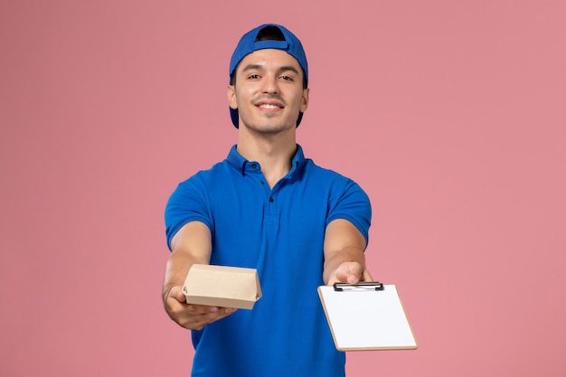 Vista frontal joven mensajero con capa uniforme azul sosteniendo un pequeño paquete de comida de entrega y un bloc de notas en la pared rosa claro