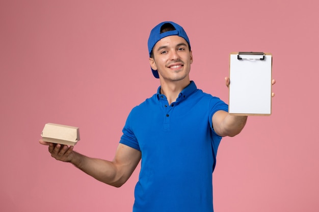 Vista frontal joven mensajero con capa uniforme azul sosteniendo un pequeño paquete de comida de entrega y un bloc de notas en la pared rosa claro