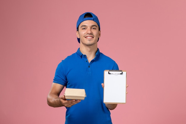 Vista frontal joven mensajero con capa uniforme azul sosteniendo un pequeño paquete de comida de entrega y un bloc de notas en la pared rosa claro