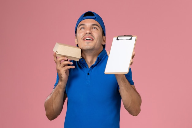 Foto gratuita vista frontal joven mensajero con capa uniforme azul sosteniendo un pequeño paquete de comida de entrega y un bloc de notas en la pared rosa claro