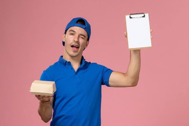 Vista frontal joven mensajero con capa uniforme azul sosteniendo un pequeño paquete de comida de entrega y un bloc de notas en la pared rosa claro